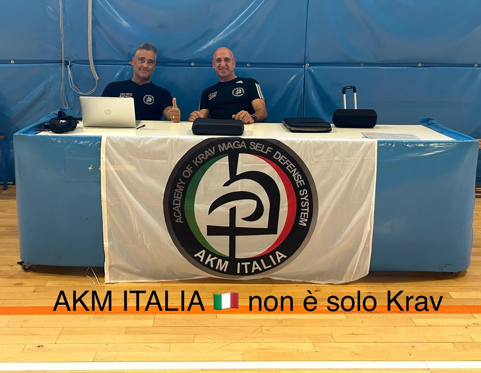 Due istruttori AKM al tavolo di un evento, davanti lo striscione di AKM Italia
